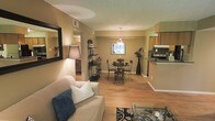 This single-story space showcases an open-concept floor plan that seamlessly connects the living, dining, and kitchen areas into one cohesive and functional environment. The living area is anchored by a comfortable seating arrangement, complemented by a decorative framed mirror and a warm floor lamp that enhance the cozy ambiance. Hardwood-style flooring extends throughout the main living spaces, adding warmth and visual continuity.

The dining area, positioned centrally within the open layout, features a round glass-and-iron table with matching chairs beneath a hanging pendant light fixture. A window behind the dining table allows natural light to filter into the space. A decorative shelving unit and tall floor vase add thoughtful styling accents throughout the room.

The kitchen, situated along one side of the open plan, is partially visible and features wood-toned cabinetry, a range, and an overhead range hood. A raised countertop creates a natural partition between the kitchen and the adjoining areas. The neutral, warm wall tones and textured ceiling tie the entire space together, contributing to an overall atmosphere that is both practical and welcoming.