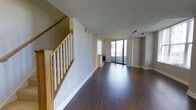This two-story residence is thoughtfully designed to balance functionality with modern aesthetics. The main level showcases rich, dark hardwood flooring that extends throughout the open-plan living area, creating a warm and inviting atmosphere. Clean neutral walls and white trim provide a timeless backdrop, while recessed and track lighting offer versatile illumination options. A standout feature of the main floor is the floor-to-ceiling sliding glass doors that open onto a private balcony, flooding the interior with natural light and offering views of the surrounding cityscape. A wet bar or kitchen prep area is visible in the background, adding to the practical appeal of the space. Connectivity between floors is provided by a well-crafted staircase featuring solid oak handrails and white-painted balusters, adding a touch of classic craftsmanship to the interior. The staircase is wide and open, contributing to the overall sense of airiness throughout. The layout is efficient yet spacious, with thoughtful architectural detailing evident in the clean lines, stepped ceiling accents, and carefully placed vents and fixtures. Overall, this residence offers a polished, move-in-ready environment well-suited for comfortable urban living.