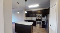 This single-floor residence offers a thoughtfully designed layout that balances modern style with practical living. The kitchen serves as a standout feature, showcasing rich dark cabinetry that extends to the ceiling for ample storage, light granite countertops, and a full suite of stainless steel appliances including a range, microwave, dishwasher, and refrigerator. A central island with matching granite provides additional workspace and a natural gathering point, while pendant lighting adds a polished finishing touch. Light wood-style flooring runs throughout the kitchen and living areas, creating a warm, cohesive aesthetic. The open living space is bright and airy, enhanced by ceiling fans and windows that invite natural light. A dedicated bedroom offers plush carpeting underfoot, a ceiling fan, and window blinds for comfort and privacy. The bathroom is finished with clean white tile flooring, a tub-shower combination, and a simple, functional layout. Overall, the space presents a clean, contemporary interior with a neutral color palette, durable finishes, and an efficient floor plan well-suited for comfortable everyday living.