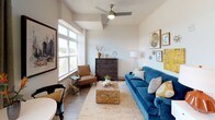 This single-floor residence offers a thoughtfully designed open-concept layout finished with light wood-style flooring throughout. The kitchen is outfitted with white shaker-style cabinetry, light quartz countertops, a grey subway tile backsplash, and a full suite of stainless steel appliances, including an electric range, built-in microwave, and dishwasher. A double undermount sink and generous counter space enhance functionality. Adjacent to the kitchen, a compact dining area accommodates a round table and seating, positioned beside a glass balcony door framed by an arched transom window. The balcony provides outdoor access with views of surrounding greenery. The living area receives abundant natural light through large windows and features a ceiling fan overhead, creating a comfortable and airy atmosphere. The bedroom includes a striking navy accent wall, a ceiling fan, and carpeted flooring, offering a cozy and stylish retreat. The bathroom is finished with white cabinetry, wood-style flooring, and clean neutral walls, maintaining the cohesive modern aesthetic found throughout the residence. A barn-style sliding door is also visible, adding a distinctive architectural detail to the interior.