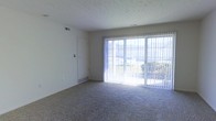 This single-floor residence offers a comfortable and functional layout designed to maximize natural light and livability. The main living area is a standout feature, showcasing expansive sliding glass doors dressed with vertical blinds that flood the room with abundant natural light and provide direct views of the surrounding greenery outdoors. The generous floor space is covered in plush wall-to-wall carpeting, adding warmth and comfort underfoot. A textured ceiling runs throughout, contributing to the room's classic residential character. Flush-mounted air vents are integrated into both the walls and floor, ensuring efficient climate control throughout the space. A clean, white interior door provides access to an adjacent room or closet, maintaining the space's simple and uncluttered aesthetic. The open layout offers flexibility for a variety of furniture arrangements, making it well-suited for everyday living. Overall, the space presents a bright, neutral, and inviting atmosphere, with its large glazed doors serving as a natural focal point that seamlessly connects the interior to the outdoor environment.