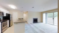 This single-floor residence showcases an open-concept design that seamlessly connects the kitchen and living areas. The living room is carpeted and centers around a classic fireplace with a simple surround, providing a focal point for the space. Large vertical blinds frame a sliding glass door that invites natural light into the room, brightening the neutral, beige-toned walls and light ceiling.

The kitchen is defined by a peninsula counter with a dark laminate surface, offering casual seating and separation from the living area. A stainless steel sink is integrated into the peninsula, and the layout allows for easy interaction between the kitchen and adjoining spaces. Recessed and flush-mount lighting overhead ensures the area is well-lit throughout.

A hallway visible from the main living space leads to additional rooms, with a bi-fold closet door indicating ample storage. HVAC vents are positioned throughout the ceiling, ensuring consistent climate control. The overall palette of soft neutrals, plush carpeting, and clean lines gives the space a calm, functional ambiance well-suited for everyday living.