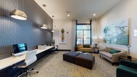 This single-floor space functions as a modern business lounge and coworking area, thoughtfully designed to balance productivity with comfort. A prominent feature is the sleek, built-in white workstation that runs along one wall, finished with a textured dark-grey accent panel and illuminated by a row of industrial-style pendant lights hanging from the elevated ceiling. Multiple computer stations with ergonomic chairs line this workspace, making it well-suited for focused tasks.

The lounge area is furnished with neutral-toned sofas and armchairs accented by teal and gold throw pillows, arranged around low circular coffee tables. A large-scale abstract painting in green, gold, and black anchors one wall, while a geometric diamond-pattern artwork adds visual interest near the workstation. The patterned carpet in grey and yellow ties the color palette together throughout the space.

Floor-to-ceiling windows with dark navy frames, horizontal blinds, and patterned drapes allow generous natural light to filter in while maintaining privacy. A glass-paneled entry door with keycard access provides a secure, professional entry point. Recessed lighting supplements the natural light, ensuring the space remains well-lit at all times. The overall ambiance is contemporary and polished, designed to support both collaborative and individual work.