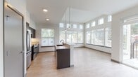 This thoughtfully designed single-floor residence showcases a clean, contemporary aesthetic with an open-concept layout that seamlessly connects the kitchen, dining, and living areas. The kitchen is a standout feature, equipped with stainless steel appliances including a refrigerator, range, and microwave, complemented by dark espresso cabinetry, a tile backsplash, and a large center island with a built-in sink and light countertop surface. Pendant lighting hangs above the island, adding both function and style. The living area benefits from dramatically tall ceilings and an expansive bank of windows, including clerestory windows positioned high on the walls, flooding the space with natural light and views of the surrounding neighborhood and greenery. Light-toned luxury vinyl plank flooring flows throughout the main living areas, lending warmth and continuity. A sliding glass door opens onto a private balcony with a metal railing, extending the living space outdoors. The bedroom features plush carpet flooring and generous windows overlooking the neighborhood. The bathroom is finished with light wood-look flooring, a tub-shower combination with tile surround, a dark-cabinetry vanity with a white undermount sink, quartz countertop, and brushed nickel fixtures and hardware throughout.