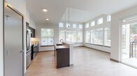 This thoughtfully designed single-floor residence showcases a clean, contemporary aesthetic with an open-concept layout that seamlessly connects the kitchen, dining, and living areas. The kitchen is a standout feature, equipped with stainless steel appliances including a refrigerator, range, and microwave, complemented by dark espresso cabinetry, a tile backsplash, and a large center island with a built-in sink and light countertop surface. Pendant lighting hangs above the island, adding both function and style. The living area benefits from dramatically tall ceilings and an expansive bank of windows, including clerestory windows positioned high on the walls, flooding the space with natural light and views of the surrounding neighborhood and greenery. Light-toned luxury vinyl plank flooring flows throughout the main living areas, lending warmth and continuity. A sliding glass door opens onto a private balcony with a metal railing, extending the living space outdoors. The bedroom features plush carpet flooring and generous windows overlooking the neighborhood. The bathroom is finished with light wood-look flooring, a tub-shower combination with tile surround, a dark-cabinetry vanity with a white undermount sink, quartz countertop, and brushed nickel fixtures and hardware throughout.