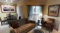 This single-floor space exudes a warm, classic charm with thoughtfully curated interiors. The living area serves as the heart of the space, anchored by a plush sofa with striped upholstery and rich leather accent pillows, complemented by a comfortable armchair. A glass-top coffee table and a decorative round tray add refined finishing touches to the seating arrangement. Crown molding along the ceiling lends an elegant architectural detail, while the warm earth-toned walls create a welcoming ambiance throughout. Natural light filters in through a window dressed with horizontal blinds and flowing sage-green curtains, brightening the room with a soft, inviting glow. A classic floor lamp beside a small round side table provides additional warmth to the corner of the room, where a vase of fresh flowers adds a lively, natural accent. Framed artwork on the walls and carefully placed decorative objects contribute to the space's cohesive and tasteful aesthetic. The overall atmosphere is one of comfort and understated elegance, making this a well-appointed space ideal for relaxed everyday living.