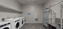 Located at 252 Avenue E in Bayonne, NJ, this shared laundry facility is designed with functionality and convenience in mind. The space is finished with large-format gray tile flooring and neutral gray walls, creating a clean and modern aesthetic throughout. Multiple front-loading washers and dryers — including both full-size and stacked units — are arranged along the walls, offering ample laundry capacity for residents. A wall-mounted digital payment and monitoring panel provides easy, tech-enabled access to machine controls and payment options, streamlining the laundry experience. Overhead wall-mounted shelving offers convenient space for laundry supplies and folded items. The facility also includes a dedicated area with heavy-duty chrome utility carts, ideal for transporting laundry and personal belongings, alongside a built-in countertop surface that provides a practical workspace. Double doors with automatic closers provide accessible entry and exit. The space is well-lit with recessed ceiling lighting and is equipped with a smoke detector and fire alarm system, ensuring safety standards are met. Overall, the facility is thoughtfully laid out to accommodate the day-to-day needs of a residential building with efficiency and ease.