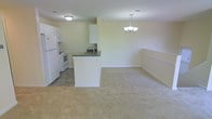 This single-floor space features an open-concept layout that integrates the kitchen and living area into one cohesive environment. The kitchen is finished with light-colored cabinetry, a dark countertop peninsula with a built-in sink, and includes white appliances — a refrigerator, stove, and range — set on tile flooring. The peninsula serves as a natural divider between the kitchen and the adjoining living area, which transitions to wall-to-wall carpeting in a warm neutral tone. The living area is generously sized, offering an open and airy feel enhanced by pale yellow walls. Overhead, a decorative chandelier with multiple light fixtures adds a touch of elegance to the dining or living space, complementing the flush-mount ceiling light in the kitchen. White chair rail molding runs along the lower portion of the walls in the living area, adding a subtle architectural detail. The overall ambiance is clean, bright, and functional, with a straightforward layout that maximizes usable floor space throughout.
