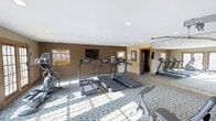 This single-floor fitness room offers a practical and well-organized space for cardiovascular and strength training. The room is furnished with multiple treadmills arranged in a row, alongside additional cardio equipment including a stair climber and a stationary bike, providing users with a range of workout options. A wall-mounted flat-screen television is positioned at the front of the room, allowing exercisers to stay entertained during their sessions.

The space features recessed ceiling lighting that ensures even illumination throughout, complemented by natural light filtering in through a window fitted with wooden blinds. The flooring is covered in a patterned carpet with a circular motif, adding a distinct visual element to the room. Deep brown accent walls contrast with the lighter ceiling, giving the space a defined, focused atmosphere.

A large mirror spans one wall, visually expanding the room and allowing users to monitor their form during workouts. A door at the rear provides additional access, while informational postings near the entrance offer guidance for users. Overall, the space is thoughtfully arranged to maximize functionality.