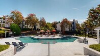 Located at 505 Riverbend Pkwy in Athens, Georgia, this single-level outdoor amenity space is designed to deliver a resort-style experience within a well-maintained residential community. The centerpiece of the space is a generously sized, freeform swimming pool with crystal-clear blue water, accessible via stainless steel handrails and surrounded by a broad, light-colored concrete pool deck. The deck is furnished with an array of chaise lounge chairs and Adirondack-style seating, ideal for relaxing under the Georgia sun. A covered cabana with a pitched shingle roof and decorative shutters sits adjacent to the pool, providing a shaded gathering spot with a bar-style counter accented in bold red detailing. String lights are strung overhead across portions of the patio, adding a warm, inviting ambiance for evening use. A dedicated outdoor dining and grilling area, outfitted with red umbrellas and casual seating, offers a social space for outdoor entertaining. The surrounding grounds are richly landscaped with mature trees displaying vibrant seasonal foliage, manicured lawns, and decorative shrubs, all enclosed by a wrought iron fence. The main clubhouse building, clad in navy blue and white siding with large windows, anchors the space architecturally and overlooks the entire pool area.