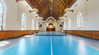 Located at 27 Park Rd in West Hartford, CT, this remarkable space is a thoughtfully adapted historic church that now serves as an indoor sports facility. The single-floor layout encompasses a generously expansive main hall, offering ample room for multiple pickleball courts laid out on a vibrant blue sport surface.

The architectural character of the original structure has been carefully preserved throughout the conversion. A dramatic Gothic-style timber vaulted ceiling soars overhead, its exposed wooden ribbing and warm tones creating a striking visual canopy above the courts. Rows of tall, arched windows line both sides of the hall, flooding the interior with abundant natural light and offering views of the surrounding West Hartford streetscape. Original stained glass windows remain intact at the ends of the hall, adding rich color and historic character to the space.

Warm wood wainscoting runs the length of the perimeter walls, complementing the original ornate woodwork near what was once the chancel area. Vintage pendant light fixtures hang from the ceiling, reinforcing the building's historic ambiance. An entry area with terrazzo flooring provides a transitional space leading into the main hall. The overall atmosphere uniquely balances architectural heritage with active recreational use.
