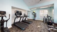 Located at 100 Rose Garden Ln in Durham, NC, this well-equipped fitness center offers a bright and energizing environment for a comprehensive workout experience. The space is characterized by its striking vaulted ceiling, which adds a sense of openness and volume, and is painted in a vibrant teal and white color scheme that creates an inviting, modern atmosphere.

Natural light pours in through a dramatic bank of large, arched windows along one wall, as well as additional windows on the side, ensuring the space feels airy and connected to the outdoors. Ceiling fans are mounted overhead to maintain comfortable airflow throughout workouts.

The fitness center is thoroughly equipped for both cardio and strength training. Multiple treadmills are positioned for easy use, accompanied by stationary and cycling bikes, and a rowing machine. A wall-mounted flat-screen television faces the cardio area for entertainment during workouts. For strength training, the space includes a multi-function cable machine, an adjustable weight bench, a full rack of dumbbells, and a selection of medicine balls and kettlebells neatly organized on a dedicated rack. The flooring features durable, patterned carpet tiles suited for a high-traffic fitness environment. A restroom is conveniently accessible directly from the fitness floor, rounding out the practical amenities of this well-appointed community fitness facility.