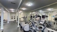 This fitness center is designed to provide a comprehensive workout environment within a single-level space. The room is outfitted with a lineup of modern cardio equipment, including treadmills, elliptical trainers, and additional machines positioned to maximize the use of the floor space. A full wall of mirrors runs along one side of the room, visually expanding the space while allowing users to monitor their form during exercise. The mirrors also reflect the overhead flush-mount lighting fixtures, which are evenly distributed across the dark ceiling to ensure consistent illumination throughout. The flooring is a durable dark rubber or vinyl material suited for high-traffic fitness use. Structural columns are integrated into the layout and painted in a neutral tone to blend with the light-colored walls. A flat-screen television is mounted and visible near the equipment area, adding an entertainment option for users. Additional free weights or benches appear to be accessible along the perimeter, rounding out the range of available workout options. The overall ambiance is clean, functional, and purposefully designed for regular physical activity.