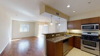 This single-story unit at 3089 James M Wood Blvd in Los Angeles offers a well-designed open-concept layout that seamlessly connects the kitchen and living areas. The kitchen is a standout feature, showcasing rich dark wood cabinetry, granite countertops with warm tonal variation, and a full suite of stainless steel appliances including a gas range, built-in microwave, dishwasher, and refrigerator. A peninsula with pendant lighting bridges the kitchen and living space, providing both additional counter space and a natural visual separator. The living and dining area is finished with warm hardwood flooring that extends throughout, enhanced by natural light from large windows and sliding glass doors fitted with vertical blinds. A chandelier anchors the dining area, adding a refined touch. The bedrooms feature plush carpeting and windows that bring in ample natural light. Two bathrooms maintain consistent design language with granite countertops and dark wood vanities; the primary bathroom offers dual sinks, a soaking tub, a separate glass-enclosed shower, and large-format tile flooring, while the secondary bathroom includes a tub-shower combination. An in-unit stacked washer and dryer adds practical convenience to the overall layout.