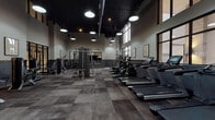 This fitness center at 1717 South Riverside Drive in Tulsa, Oklahoma, offers a spacious, single-floor layout designed with a modern industrial aesthetic. Soaring ceilings with exposed ductwork and dark pendant lighting create a bold, contemporary atmosphere throughout the facility. The flooring features a checkered pattern of charcoal and slate-toned tiles, adding visual texture while providing a durable, functional surface.

Large steel-framed windows line the perimeter, allowing ample natural light to fill the space while providing views of the surrounding exterior. The two-toned walls, featuring a deep gray lower half accented by warm wood trim, complement the overall industrial design. Motivational framing and artwork are displayed along the walls, enhancing the workout atmosphere.

The cardio zone is well-stocked with a row of Spirit treadmills equipped with individual screens, alongside ellipticals and recumbent bikes. The strength training area includes multiple selectorized weight machines, a dual cable functional trainer, a full dumbbell rack, and a large wood-framed mirror with a mounted flat-screen television. Black-framed glass partitions offer partial sightlines into adjacent spaces, contributing to an open, connected feel throughout the facility.