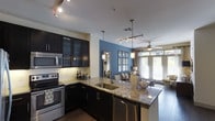 This single-floor space showcases a thoughtfully designed open-concept layout that seamlessly connects the kitchen and living area. The kitchen is a standout feature, boasting dark espresso cabinetry with glass-front upper cabinets, granite countertops, and a large kitchen island with an undermount sink — ideal for both cooking and entertaining. Stainless steel appliances, including a range, microwave, and dishwasher, complement the sleek aesthetic. Under-cabinet lighting adds warmth and functionality to the workspace. A bold blue accent wall anchors the transition between the kitchen and the living area, adding a striking pop of color to the neutral palette. Pendant lights and a track lighting system overhead provide layered illumination throughout. The living area is bright and inviting, benefiting from large windows dressed with floor-length curtains that allow generous natural light to filter in. A ceiling fan enhances comfort in the space. Dark hardwood-style flooring runs throughout, tying together the modern design elements cohesively. Overall, the space exudes a contemporary and sophisticated ambiance, balancing bold design choices with practical, well-appointed features.