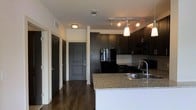 This single-floor space showcases a contemporary design anchored by a well-appointed kitchen. Dark espresso cabinetry extends from floor to ceiling, providing ample storage, while granite countertops add a polished, upscale feel. Black appliances, including a refrigerator and built-in microwave, contribute to the kitchen's cohesive, modern aesthetic. A peninsula with a stainless steel sink serves as both a functional workspace and a natural divider between the kitchen and the adjacent living area. Pendant lighting hangs above the peninsula, while a track lighting system illuminates the kitchen workspace. A neutral-toned tile backsplash adds texture and contrast against the dark cabinetry. Rich, dark hardwood flooring runs throughout the space, creating a warm and unified look. The hallway leading from the kitchen features multiple doors, likely accessing bedrooms, closets, and a main entry, all finished in the same dark tone as the cabinetry for a consistent design language. The overall ambiance is sleek and modern, with a carefully coordinated color palette of dark neutrals and warm wood tones.