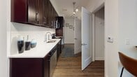 This single-floor studio unit offers a thoughtfully organized open-concept layout that seamlessly integrates the sleeping, living, and kitchen areas into one cohesive space. Rich hardwood-style flooring flows throughout, anchoring the modern aesthetic and providing visual continuity.

The sleeping and living area is generously proportioned, accommodating a full-sized bed alongside lounge seating and a compact dining arrangement. A large window allows abundant natural light to fill the space, offering views of surrounding greenery. Crisp white walls and clean lines contribute to an airy, contemporary feel.

The kitchen runs along one wall in a galley-style configuration, featuring dark espresso cabinetry contrasted by white quartz countertops and a subway tile backsplash. Stainless steel appliances, including a refrigerator, range, microwave, and dishwasher, are integrated throughout. Track lighting overhead ensures the workspace is well-illuminated.

A hallway connects the kitchen to a walk-in closet and a full bathroom. The bathroom is finished with light tile flooring, a dark vanity with ample storage, a wide mirror, and a tub-shower combination. The overall design balances function and style in a compact, efficient footprint.