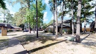 This single-story residence presents a classic suburban character defined by its brick exterior and a dark, pitched roof with what appears to be a chimney detail. The home is set well back from the street, framed by a striking collection of tall, mature pine and deciduous trees that provide generous natural shade across the expansive front yard. The grounds feature a largely open lawn area with a mix of grass and natural ground cover, lending the property a relaxed, established feel. A driveway leads toward what appears to be an attached garage, offering practical convenience. The surrounding neighborhood, visible in the background, reflects a similarly mature, tree-lined character with comparable residential homes. The overall ambiance of the exterior is one of quiet, established suburban living, with the dense tree canopy creating a sense of privacy and seclusion despite its proximity to the street. The natural landscaping, combined with the traditional brick construction, gives the home a grounded, timeless appearance well-suited to its verdant, wooded surroundings.
