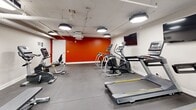 Located at 70 Spadina Rd in Toronto's Annex neighbourhood, this fitness facility offers a well-organized and fully equipped exercise room designed to meet a variety of workout needs. The space features a practical, no-frills layout with durable rubber flooring throughout, providing a stable and comfortable surface for exercise. Overhead, bold octagonal and rectangular light fixtures ensure the room is brightly and evenly illuminated.

The cardio section is a focal point of the gym, housing multiple treadmills, elliptical trainers, and stationary bikes — both upright and recumbent — catering to users of varying fitness preferences. A dedicated free weights area includes a full rack of dumbbells in graduated sizes, accompanied by a flat bench, making it suitable for strength training. Wall-mounted mirrors along one side of the room enhance the sense of space and allow users to monitor their form during workouts. Two flat-screen televisions are mounted on the walls, adding an entertainment element to longer cardio sessions.

The room's design is clean and utilitarian, with white walls accented by a striking orange feature wall that adds a bold, energetic visual contrast. Exposed ceiling infrastructure, including visible pipes and ductwork, gives the space an industrial character. The facility is compact yet efficiently arranged to maximize usability within its footprint.