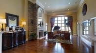 This single-story space exudes classic elegance through a carefully curated blend of architectural details and refined finishes. The centerpiece of the room is a striking floor-to-ceiling stone column framing a grand arched fireplace with an ornate white mantle, anchoring the space with a sense of timeless character. Rich, wide-plank hardwood floors extend throughout, reflecting the warm glow of recessed ceiling lights and a classic table lamp, enhancing the inviting atmosphere. Crown molding runs along the upper walls, adding a polished architectural finish. A large arched window dressed in dramatic red and green drapery floods the space with natural light, drawing the eye outward. The seating area features traditional-style upholstered armchairs and a sofa arranged around a central coffee table atop a patterned area rug, creating an intimate yet open gathering space. Lush potted plants are positioned throughout, softening the formal elements with natural texture. The warm golden wall tones complement the wood and stone materials, resulting in a cohesive, sophisticated interior with a welcoming, traditional sensibility.