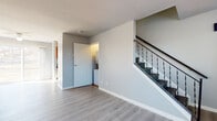 This two-floor space is designed with a clean, neutral aesthetic that emphasizes functionality and simplicity. The main level features light grey laminate flooring that runs throughout the open living areas, complemented by soft grey walls and crisp white trim and baseboards. A staircase with a dark metal balustrade connects the two floors, adding a subtle contrast to the otherwise light interior. The main level includes a spacious living area with multiple windows that allow natural light to fill the room, as well as a dedicated laundry area accessible through a paneled interior door. The kitchen is equipped with warm-toned wooden cabinetry, granite-look countertops, a subway tile backsplash, and stainless steel and white appliances, including a refrigerator and electric range. The upper level transitions to carpeted bedrooms, each featuring sliding panel closets and baseboard heating. A full bathroom on this level includes a tiled tub-surround, grey floor tiles, and a window with a privacy blind. Baseboard heating units are present throughout both levels, ensuring consistent climate control. The overall ambiance is understated and move-in ready, with a cohesive neutral palette throughout.