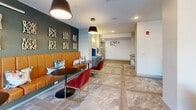 Located at 10 Calm Water Drive in Asheville, NC, this single-floor community lounge space is thoughtfully designed to offer both comfort and visual sophistication. The space is unified by large-format marble-look porcelain tile flooring that flows seamlessly throughout, creating a cohesive and polished foundation.

The living area anchors one end of the space, bathed in abundant natural light streaming through expansive floor-to-ceiling glass windows and a glass-panel garage-style door that frames views of the surrounding exterior. A striking sputnik-style chandelier serves as a focal point overhead, complementing the wall-mounted flat-screen television. Plush, neutral-toned lounge chairs are arranged around a sleek coffee table, creating an inviting gathering spot. Vibrant, large-scale watercolor abstract artwork adorns the white walls, adding a dynamic artistic element.

The dining area transitions naturally from the lounge, featuring a long banquette upholstered in warm cognac leather set against a deep teal accent wall decorated with sculptural wall art. Red velvet accent chairs and round bistro tables provide flexible seating. A compact high-top table with upholstered counter stools offers an intimate secondary dining option, surrounded by additional large framed abstract paintings. A service counter with cabinetry is accessible nearby, rounding out the functional amenities of this well-appointed community space.