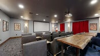This single-floor entertainment space is designed as a private theater and lounge, combining cinematic function with warm, rustic character. The screening room is outfitted with rows of generously sized leather chairs arranged to face a large projection screen, served by a ceiling-mounted projector overhead. Dark painted ceilings enhance the theatrical atmosphere, while recessed lighting and decorative wall sconces provide ambient illumination. Bold red curtains frame the screen wall and accent the entry, adding a classic cinema feel. Distressed wood pillar accents and patterned carpet in a muted gray-and-blue tone lend texture throughout.

Adjacent to the screening area is a flexible lounge and gathering space featuring live-edge wood communal tables, upholstered blue chairs, and bistro-height seating arrangements suited for social events or informal meetings. A built-in cabinet with a granite countertop and a striking deep-blue handcrafted tile backsplash serves as a concession-style station, complete with a classic popcorn machine. Framed movie posters decorate the walls, reinforcing the cinematic theme. The space opens toward a larger dining or common area beyond, distinguished by a stone fireplace, exposed beams, and tall windows that introduce natural light into the broader facility.