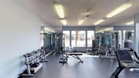 Located at 955 Airport Rd in Destin, Florida, this fitness center offers a well-appointed, single-floor workout facility designed to support a comprehensive exercise experience. The space is outfitted with a wide selection of equipment, including a full dumbbell rack with varying weights, an adjustable workout bench, a cable machine, and a multi-station strength training unit, catering to both free weight and machine-based training. Large floor-to-ceiling mirrors line the walls, enhancing the sense of openness and allowing users to monitor their form during workouts. The flooring consists of durable rubber or rubberized tile matting, ideal for heavy equipment use and providing safety underfoot. Natural light filters in through a set of multi-pane windows that frame an outdoor view of palm trees and open grounds, creating a bright and pleasant atmosphere. Overhead fluorescent lighting supplements the natural light, ensuring the space remains well-lit throughout the day. A ceiling fan provides additional air circulation, contributing to a comfortable workout environment. The overall aesthetic is clean and functional, with a neutral gray and white color palette that gives the facility a modern, professional appearance.