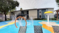 This residential community is designed to offer both functional amenities and a welcoming atmosphere. The outdoor area is a standout feature, showcasing a colorful splash pad and pool zone equipped with water jets, an arched orange water feature, and lounge chairs positioned on a bright, wave-patterned surface. Surrounded by iron fencing and mature trees, the space provides a lively yet secure environment ideal for recreation and relaxation. The multi-story building visible in the backdrop features balconies with railings, contributing to the community's cohesive architectural character.

Inside, the mail center lobby presents a clean, contemporary aesthetic with soft blue-gray walls, light tile flooring, and a dark ceiling that creates a stylish contrast. Large floor-to-ceiling windows flood the space with natural light and offer views of the surrounding greenery. Multiple banks of bronze and brushed-metal mailboxes line both sides of a central island, which includes a built-in parcel drop slot for convenient package handling. Framed geometric artwork adorns the walls, adding a curated, modern touch. Recessed lighting and wall-mounted fixtures further enhance the polished, well-maintained ambiance of this communal space.