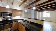 This single-floor space blends historic architectural details with modern updates to create a distinctive and functional living environment. A central hallway serves as the spine of the unit, featuring rich, wide-plank hardwood floors that run throughout and overhead exposed dark wood beams that lend a sense of industrial charm and height to the interior. A pendant light fixture hangs along the corridor, while a glass block panel embedded in the wall introduces diffused natural light without sacrificing privacy — a thoughtful architectural detail that adds character to the passage.

The kitchen is a standout feature, showcasing dark granite countertops that wrap around the perimeter and contrast cleanly with light maple cabinetry. A double-basin stainless steel sink anchors the workspace, and overhead pendant lights echo the warm tones of the exposed beam ceiling above. Open shelving adds a practical, airy touch, while a pass-through window connects the kitchen visually to the adjacent living area, enhancing the open flow of the space. The continuation of exposed beams throughout ties all areas together, reinforcing the space's cohesive, loft-inspired aesthetic.