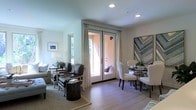 This single-story space presents an open-concept floor plan that seamlessly integrates the living and dining areas into one cohesive, functional environment. The living area is anchored by a neutral-toned area rug and furnished with a mix of leather and upholstered seating, complemented by a small ottoman coffee table. A framed landscape artwork and a classic table lamp add warmth and personality to the space.

The dining area features a round table surrounded by elegant upholstered chairs with nailhead trim detailing, positioned beneath a bold geometric wall art piece that serves as a striking focal point. Light hardwood flooring extends throughout both areas, creating visual continuity and a clean, modern aesthetic.

A set of French doors with white curtains serves as both a design element and a functional gateway to an outdoor space, filling the interior with natural light and offering a direct connection to the exterior. Recessed ceiling lighting provides even illumination throughout. The overall ambiance is warm, refined, and inviting, with a carefully curated blend of traditional and contemporary furnishings.