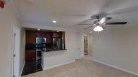 This single-floor residence offers a thoughtfully designed open-concept layout that seamlessly connects the kitchen and living areas. The kitchen is a standout feature, showcasing rich dark wood cabinetry that extends to the ceiling, complemented by dark granite countertops and a peninsula that provides additional workspace and casual seating. Stainless steel appliances — including a French door refrigerator with ice and water dispenser, a range with electric cooktop, and an over-the-range microwave — complete the modern kitchen ensemble. The dark tile flooring in the kitchen provides a striking contrast to the warm, neutral carpet that flows throughout the living area.

Crown molding runs along the ceiling, adding a refined architectural detail that elevates the overall aesthetic. Recessed lighting keeps the space bright, while a ceiling fan with integrated light fixture in the living area enhances both comfort and illumination. The neutral, light-toned walls create a clean and versatile backdrop throughout. A hallway visible from the main living area leads to additional rooms, including what appears to be a bathroom with tile flooring. White louvered doors add a classic touch to the interior design. Overall, the space presents a balanced combination of functional design and polished finishes.