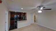 This single-floor residence offers a thoughtfully designed open-concept layout that seamlessly connects the kitchen and living areas. The kitchen is a standout feature, showcasing rich dark wood cabinetry that extends to the ceiling, complemented by dark granite countertops and a peninsula that provides additional workspace and casual seating. Stainless steel appliances — including a French door refrigerator with ice and water dispenser, a range with electric cooktop, and an over-the-range microwave — complete the modern kitchen ensemble. The dark tile flooring in the kitchen provides a striking contrast to the warm, neutral carpet that flows throughout the living area.

Crown molding runs along the ceiling, adding a refined architectural detail that elevates the overall aesthetic. Recessed lighting keeps the space bright, while a ceiling fan with integrated light fixture in the living area enhances both comfort and illumination. The neutral, light-toned walls create a clean and versatile backdrop throughout. A hallway visible from the main living area leads to additional rooms, including what appears to be a bathroom with tile flooring. White louvered doors add a classic touch to the interior design. Overall, the space presents a balanced combination of functional design and polished finishes.