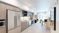 Located in Portland's vibrant Northwest District at 1985 NW Savier St, this well-designed one-bedroom, one-bathroom residence offers a smart, efficient layout across a single floor. Light wood-style flooring flows throughout, lending warmth and visual continuity to the space.

The open-plan kitchen and living area forms the heart of the unit. The kitchen is outfitted with stainless steel appliances—including a French door refrigerator, gas range, and dishwasher—paired with two-tone cabinetry in soft sage and natural wood tones, a tile backsplash, and under-cabinet lighting. Open shelving adds both function and style.

The living room receives generous natural light through large windows overlooking the surrounding neighborhood. A hallway with recessed lighting connects the main living area to the private bedroom and bathroom, with a closet providing additional storage.

The bedroom features a geometric accent wall and ample space for a full furniture arrangement. The bathroom offers a clean, modern aesthetic with sage-green walls, a tiled tub-shower combo, a floating vanity with a white countertop, and a large illuminated mirror.

A dedicated hallway with a wall-mounted thermostat and access to multiple rooms ensures a logical, practical flow throughout this thoughtfully designed urban residence.