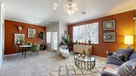 This thoughtfully designed single-story, one-bedroom, one-bathroom residence offers a warm and eclectic aesthetic anchored by striking terracotta and burnt-orange accent walls throughout. The open living and dining area is bathed in natural light from large windows with vertical blinds, and features plush carpet flooring, crown molding, and a ceiling fan. The living space provides a comfortable layout with ample room for seating, while a dedicated dining area offers built-in shelving and direct access to the outdoors. The kitchen is equipped with granite-style countertops, a full suite of white appliances including a refrigerator, range, and overhead microwave, complemented by hardwood-style vinyl flooring and generous cabinet storage. A convenient in-unit laundry area accommodates a full-size washer and dryer. The spacious primary bedroom features soft carpet, a ceiling fan, and a large window that invites natural light. The bathroom includes a wide vanity with granite-style countertops and ample storage. Throughout the residence, warm earth tones and cohesive design details create an inviting, desert-inspired ambiance that is both functional and full of character.