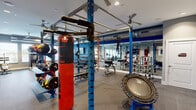 This expansive fitness and recreation facility is thoughtfully designed to accommodate a wide range of activities across multiple interconnected zones. The strength training area is anchored by a bold blue power rack system complete with free weights, benches, resistance bands, a heavy punching bag, and a mini trampoline, all set on durable rubber flooring beneath exposed industrial-style ductwork and ceiling fans. Full-length mirrors line the walls, enhancing the sense of space throughout.

A dedicated cardio zone features rows of treadmills set against a vivid blue accent wall, while a separate cycling studio offers spin bikes, yoga mats, foam rollers, and wall-mounted televisions for guided workouts. Natural light filters in through multiple windows fitted with blinds, keeping the environment bright and energizing. Ceiling fans throughout ensure comfortable airflow during intense sessions.

The facility also includes a well-equipped cardio and stretching lounge with recumbent bikes and elliptical machines arranged along a long, open floor plan. Adjacent to the fitness areas, a stylish game room provides a welcoming retreat, featuring a pool table, shuffleboard, electronic dartboards, classic arcade cabinets, a pinball machine, and a reclaimed wood accent wall. An elegantly appointed corridor with decorative iron railings connects the various spaces, completing this well-rounded amenity facility.