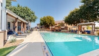 Located at 3650 Custer Pkwy in Richardson, Texas, this outdoor swimming pool area is a well-appointed recreational amenity designed for relaxation and leisure. The centerpiece is a generously sized, crystal-clear swimming pool with a sparkling turquoise finish, accessible via built-in entry steps with stainless steel handrails. The pool is surrounded by a broad, light-colored concrete sun deck that provides ample space for lounging and movement.

Numerous blue lounge chairs are arranged along the poolside, offering residents dedicated spaces to sunbathe and unwind. Multiple covered pergola structures, constructed with warm wood tones and brick columns, provide shaded retreats and are furnished with additional seating and patio tables. Patio umbrellas are strategically positioned around the deck to offer further shade options throughout the day.

Mature trees frame the perimeter of the pool area, contributing natural shade and a sense of privacy. Ornamental grasses and native landscaping plantings line the surrounding walkways, enhancing the aesthetic appeal. The adjacent brick residential buildings, featuring balconies and large windows, overlook the pool area and create an intimate, community-oriented atmosphere. The overall setting is open-air, bathed in abundant natural Texas sunlight, making it an inviting outdoor destination for warm-weather enjoyment.