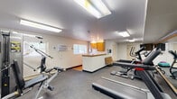 Located at 20 Jordan Dr in Whitehall, PA, this single-floor exercise room offers a well-equipped fitness facility designed to support a variety of workout routines. The space is laid out efficiently, with gray carpet covering the main exercise area and smooth transitions between workout zones.

The cardio section features multiple treadmills, a stationary bike, and an elliptical trainer, all positioned alongside a large wall-mounted mirror that enhances the sense of space and allows users to monitor their form. A multi-function cable and weight machine provides strength training options, while a dedicated free weights area includes dumbbells and step platforms for versatile training. A wall-mounted flat-screen TV is positioned for convenient viewing during workouts, and a wooden bench provides a seating area for rest or stretching.

Bright fluorescent lighting illuminates the room from above, and windows along one wall allow natural light to filter in. A wall-mounted air conditioning unit helps maintain a comfortable environment. A small kitchenette area with wooden cabinetry and a countertop is tucked into one corner, adding a practical amenity to the space. Two entry doors provide easy access. Overall, the facility is clean, functional, and thoughtfully organized to accommodate a range of fitness activities.