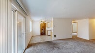 Located at 12514 SE 7th St in Vancouver, Washington, this one-bedroom, one-bathroom unit offers a practical and well-organized single-floor layout. The galley-style kitchen is efficiently designed with oak-trimmed white cabinetry lining both walls, providing ample storage space. White appliances — including an electric range with overhead microwave and a dishwasher — complement the light countertops and tile flooring, creating a clean, functional workspace. A hallway leads from the kitchen toward the remaining rooms, maintaining a logical flow throughout the unit. The generously sized primary bedroom features plush wall-to-wall carpeting, a wide sliding mirrored closet, and a window overlooking greenery outside, allowing natural light to fill the room. An additional carpeted room with a standard closet offers flexible use as a secondary living or storage space. The bathroom is bright and clean, featuring a wide vanity with oak cabinetry, a large mirror, skylight-style overhead lighting, and tile flooring. A dedicated utility room adds everyday convenience. Throughout the unit, neutral white walls and consistent finishes create a cohesive and move-in ready environment suited for comfortable living.