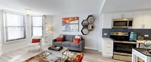 This well-appointed single-floor space showcases a thoughtfully designed open-concept layout that seamlessly connects the living area and kitchen. The living space features light hardwood-style flooring that runs throughout, contributing to a bright and airy atmosphere. Large windows dressed with horizontal blinds allow ample natural light to fill the room, enhancing the clean, modern aesthetic.

The living area is anchored by a contemporary gray sofa accented with rust-orange throw pillows, paired with a sleek glass-and-metal coffee table. A vibrant, multicolored area rug adds warmth and character underfoot, while decorative elements — including a bold, colorful wall painting and a cluster of hexagonal mirrors — lend a curated, artistic touch to the space.

The kitchen, visible from the living area, features crisp white cabinetry, dark slate-toned tile backsplash, and dark countertops, creating a striking contrast that feels both modern and functional. Stainless steel appliances complete the kitchen's polished look.

The space benefits from smooth white ceilings and neutral wall tones, providing a versatile backdrop that balances the vibrant decor. Overall, the unit offers a cohesive blend of contemporary design and comfortable living in a compact, efficiently organized floor plan.