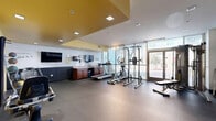 Located at 45 E 12th St in National City, California, this fitness center offers a comprehensive exercise environment within a single, efficiently designed floor. The space is flooded with natural light through floor-to-ceiling glass panels and double glass entry doors that face the street, creating an open and energizing atmosphere. Rubber flooring throughout provides durability and safety for a variety of workout activities.

The exercise area is equipped with a diverse selection of cardio and strength training machines, including an elliptical trainer, a recumbent bike, a cable/pulley functional trainer, and a multi-station power rack. A dedicated stretching and mat workout zone is defined by a wall-mounted exercise mat, complemented by a medicine ball rack stocked with multiple weighted balls. Dumbbells are available along one wall, offering a complete free weights section.

A large wall mirror expands the sense of space while allowing users to monitor their form. Motivational wall decals and signage add an encouraging atmosphere throughout. Recessed and track lighting illuminate the interior evenly. A built-in counter with a sink and cabinetry provides a convenient utility station. A wall-mounted television serves as an additional amenity. The space also connects to an adjacent hallway for easy access to surrounding facilities.