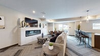 The Clubroom at 415 Hickory Dr in Cannon Falls, MN is a well-appointed single-floor community gathering space designed with modern sensibility and practical functionality. The open-concept layout seamlessly connects the living, dining, and kitchen areas, creating a welcoming environment for residents and guests alike.

The living area serves as the centerpiece of the space, anchored by a sleek linear fireplace framed in white tile with a wall-mounted flat-screen television above. Herringbone-patterned carpet tiles extend throughout, adding visual texture and warmth to the neutral color palette. Recessed lighting illuminates the space throughout.

The full kitchen is equipped with stainless steel appliances, including a full-size refrigerator and range, complemented by rich dark wood cabinetry and light granite countertops. Light hardwood-style flooring runs through the kitchen area, and a built-in water fountain station adds a practical amenity.

The dining area accommodates multiple seating configurations, including a formal dining table with upholstered chairs and a bar-height counter seating area. Large windows and a glass door provide abundant natural light and direct access to an outdoor patio with views of the surrounding landscape. A set of glass double doors at the entry connects the clubroom to the broader building, completing this versatile and elegantly designed community space.