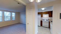 This single-floor residential space offers a practical and comfortable layout designed for everyday living. The living area features soft wall-to-wall carpeting underfoot and neutral-toned walls that create a calm, welcoming atmosphere. A window allows natural light to filter into the room, brightening the space and providing a connection to the outdoors. Adjacent to the living area, a paneled interior door leads to additional spaces within the unit. The kitchen is well-appointed with warm wood cabinetry that provides ample storage, complemented by white appliances including a gas range and a full-size refrigerator. The kitchen floor features light-toned tile, offering a clean contrast to the carpeted living area. A skylight or flush ceiling fixture overhead keeps the kitchen bright and functional. The overall layout is efficient and cohesive, with a smooth flow between rooms. Neutral color palettes throughout lend the space a timeless, move-in-ready quality. This unit is well-suited for those seeking a straightforward, functional living environment with classic residential finishes.