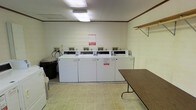 This shared laundry facility is a straightforward, single-floor space designed for practical use. A row of five coin-operated top-load washing machines lines the back wall, each equipped with individual coin-payment consoles. Posted notices and instructions are displayed on the machines for user reference. The light-colored walls are accented with a dark wood-tone crown molding trim near the ceiling, lending a modest decorative touch to the otherwise utilitarian space. Overhead, fluorescent lighting provides bright, even illumination throughout the room. A wall-mounted shelf on the right side offers convenient storage for laundry supplies, while a large folding table positioned along the right wall provides ample space for sorting and folding clothes. A trash bin is accessible near the machines, supporting cleanliness and ease of use. The flooring consists of durable, light-toned vinyl tile that is easy to maintain. The overall layout is efficient and organized, prioritizing functionality with clear circulation space between the machines and the folding area.
