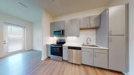 Located at 1251 Ray Charles Blvd in Tampa, FL, this single-floor residence offers a thoughtfully designed layout with modern finishes throughout. The kitchen is a functional highlight, featuring stainless steel appliances including a range, over-the-range microwave, and dishwasher, complemented by sleek grey cabinetry, white countertops, and a classic subway tile backsplash. Light wood-tone flooring runs continuously throughout the space, lending warmth and visual cohesion to every room. The open living area connects seamlessly to the kitchen, and a stylish barn-style sliding door adds a contemporary architectural touch while providing access to a closet with built-in shelving. The bedroom is bright and airy, benefiting from a generous bank of windows with horizontal blinds that allow for natural light control, along with a ceiling fan for added comfort. The bathroom is clean and well-appointed, featuring a tiled soaking tub and shower combination with a curved rod, wood-look flooring, and a simple, neutral palette. A corridor connects the kitchen to the entry, with the full-size refrigerator positioned conveniently near the entrance. The overall ambiance is modern, light-filled, and move-in ready.