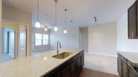 This single-floor space showcases an open-concept layout that integrates the kitchen and living areas into one cohesive environment. The kitchen is anchored by a central island topped with light granite countertops, housing a stainless steel undermount sink paired with a brushed gold pull-down faucet. Dark espresso cabinetry with brushed nickel hardware runs beneath the island, providing ample storage. Three pendant lights hang above the island, offering focused task lighting while contributing to the space's modern aesthetic.

The kitchen flows naturally into the adjoining living area, which features plush carpeting that contrasts with the hardwood flooring of the kitchen. A ceiling fan with integrated lighting serves the living space, and a window along the far wall allows natural light to filter in, brightening the neutral, light gray walls throughout.

Track lighting is also present near a hallway leading to additional rooms, enhancing overall illumination. The clean lines, neutral color palette, and thoughtful lighting choices throughout create a bright, functional, and welcoming atmosphere suited to everyday living.