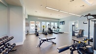 This fitness center is thoughtfully designed across two dedicated exercise rooms, offering a comprehensive workout experience. The main exercise area features high ceilings with modern linear pendant lighting and soft blue-gray walls that create a calm, motivating atmosphere. Durable speckled rubber flooring runs throughout, providing both safety and comfort underfoot. Large windows line the walls, flooding the space with natural light and offering views of the surrounding outdoor amenity areas. This primary room is outfitted with a robust selection of cardio equipment, including treadmills, ellipticals, and a stair climber, as well as strength training machines, free-weight benches, a dumbbell rack, and a full cable and multi-press rack system. Wall-mounted flat-screen TVs enhance the workout experience. A separate, dedicated studio room provides a quieter setting for focused training, equipped with a spin bike and a rowing machine, along with ample natural light from tall portrait-style windows. A mirror wall in the main area expands the sense of space, while direct-access exterior doors allow for easy entry and exit. The overall design is clean, modern, and functional, catering to a wide range of fitness routines.