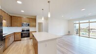 This well-designed two-bedroom, two-bathroom residence in Menomonee Falls, WI offers a thoughtfully laid-out single-floor plan with a clean, modern aesthetic throughout. The open-concept kitchen and living area serves as the heart of the home, showcasing warm medium-toned wood cabinetry, a large center island with pendant lighting, white quartz countertops, a subway tile backsplash, and a full suite of stainless steel appliances. Light hardwood flooring flows seamlessly through the kitchen and living room, where large sliding glass doors and a wide transom window flood the space with natural light and provide direct access to a private balcony with outdoor views. Both bedrooms are carpeted and feature ceiling fans, with the generously sized primary bedroom offering its own closet space. A dedicated walk-in closet with built-in shelving provides ample storage. The two bathrooms are finished with warm wood vanities and white countertops; one includes a dual-sink vanity and glass-enclosed shower, while the other features a combination tub and shower. The neutral, light-filled palette and quality finishes throughout create a comfortable and welcoming living environment.