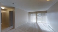 This single-floor space features a spacious, open living area characterized by neutral tones and a clean, understated aesthetic. The room is finished with wall-to-wall plush carpeting in a light beige tone, contributing to a soft and comfortable atmosphere underfoot. A textured popcorn ceiling adds a classic touch overhead, while the crisp white walls provide a blank canvas adaptable to a variety of interior styles.

A standout feature of the living area is the expansive floor-to-ceiling sliding glass door, dressed with vertical blinds that allow for adjustable light control. This large glazed opening floods the room with generous natural light, casting bright streaks across the carpet and creating an airy, open feel. The sliding door provides direct access to an outdoor area, where glimpses of greenery are visible through the blinds.

The layout of the living area is open and versatile, offering ample room for a range of furniture arrangements. A doorway visible on the left side of the space leads to an adjacent room, suggesting a functional floor plan designed for ease of movement. Overall, the space presents a bright, neutral, and move-in-ready environment.